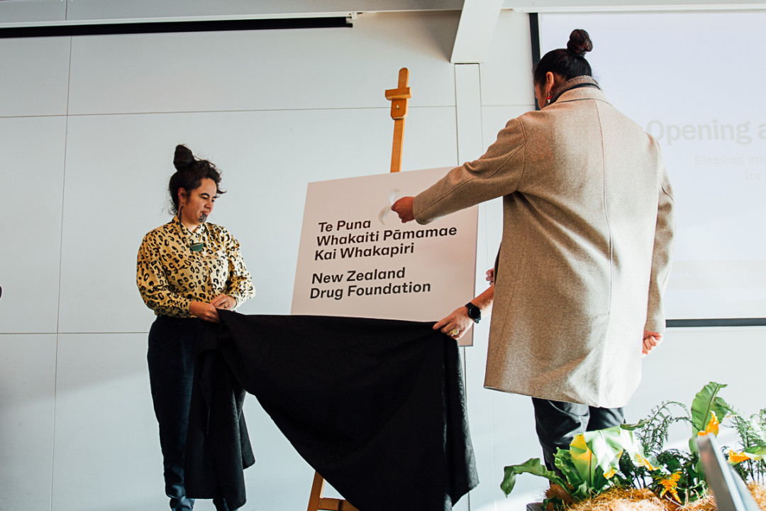 Māori woman and man pulling black cloth of easel with sign which says "Te Puna Whakaiti Pāmamae Kai Whapakiri - New Zealand Drug Foundation"