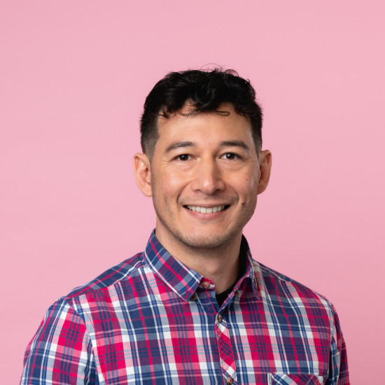 Man wearing a checked red and blue shirt, smiling at the camera in front of a pink background. 